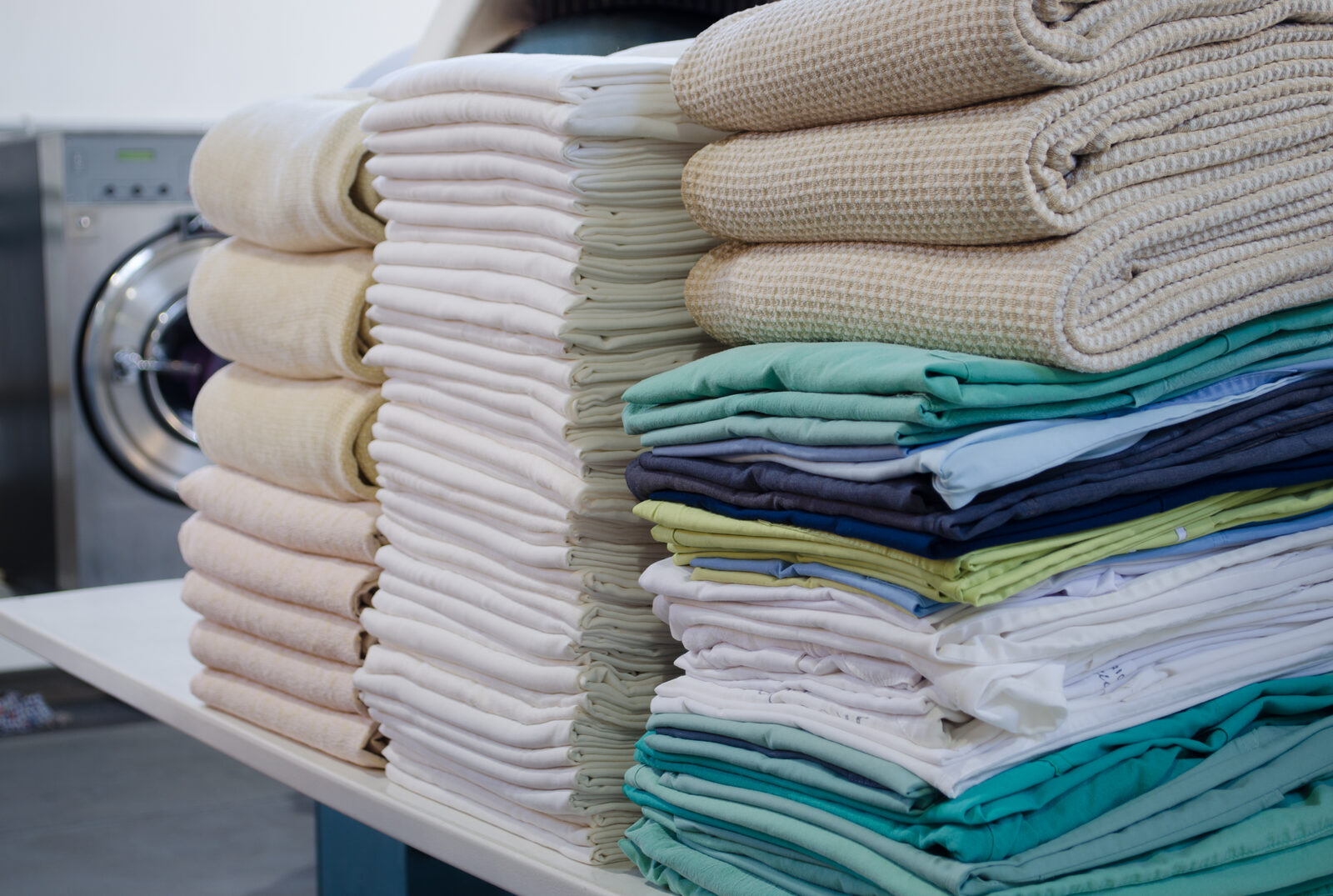 Des piles de linge repassé et plié, serviettes et draps, devant une machine à laver